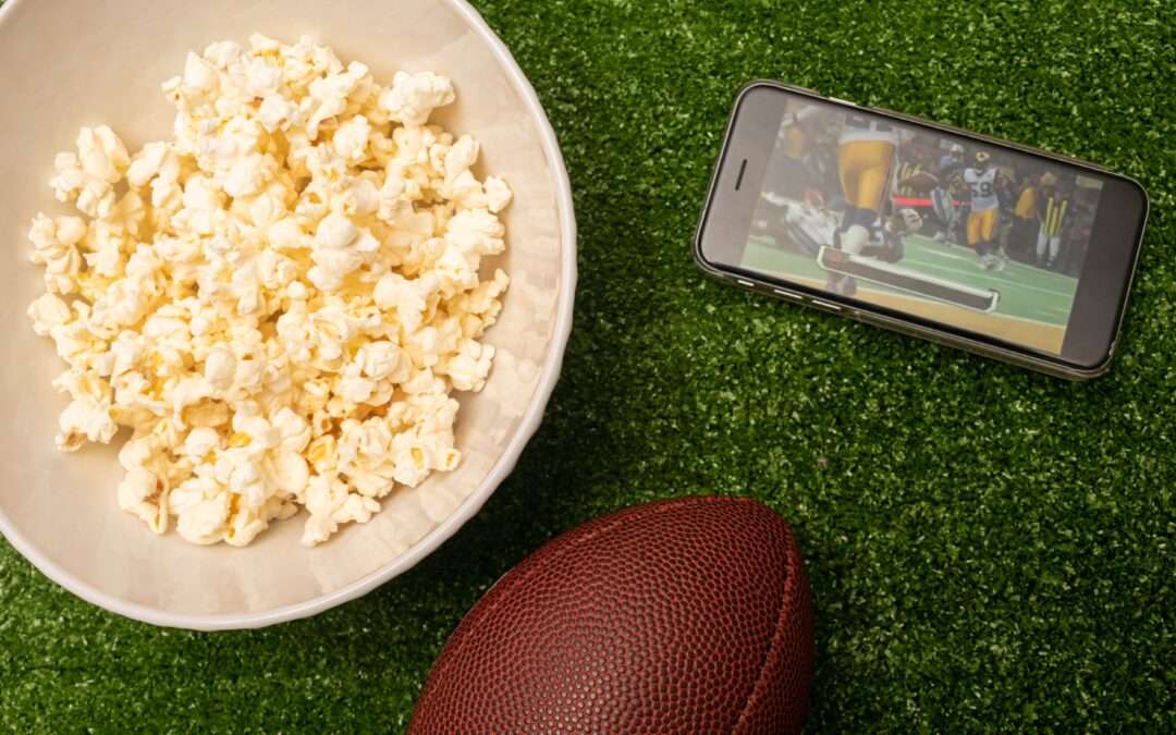 comfortable, Super bowl watch party, American football flat lay photography with popcorn, beer, american football ball and cell phone with american football game on green grass